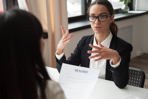 Confident millennial female applicant in glasses talking at job interview answering questions, vacancy candidate making first impression concept and introducing herself speaking to hr holding resume