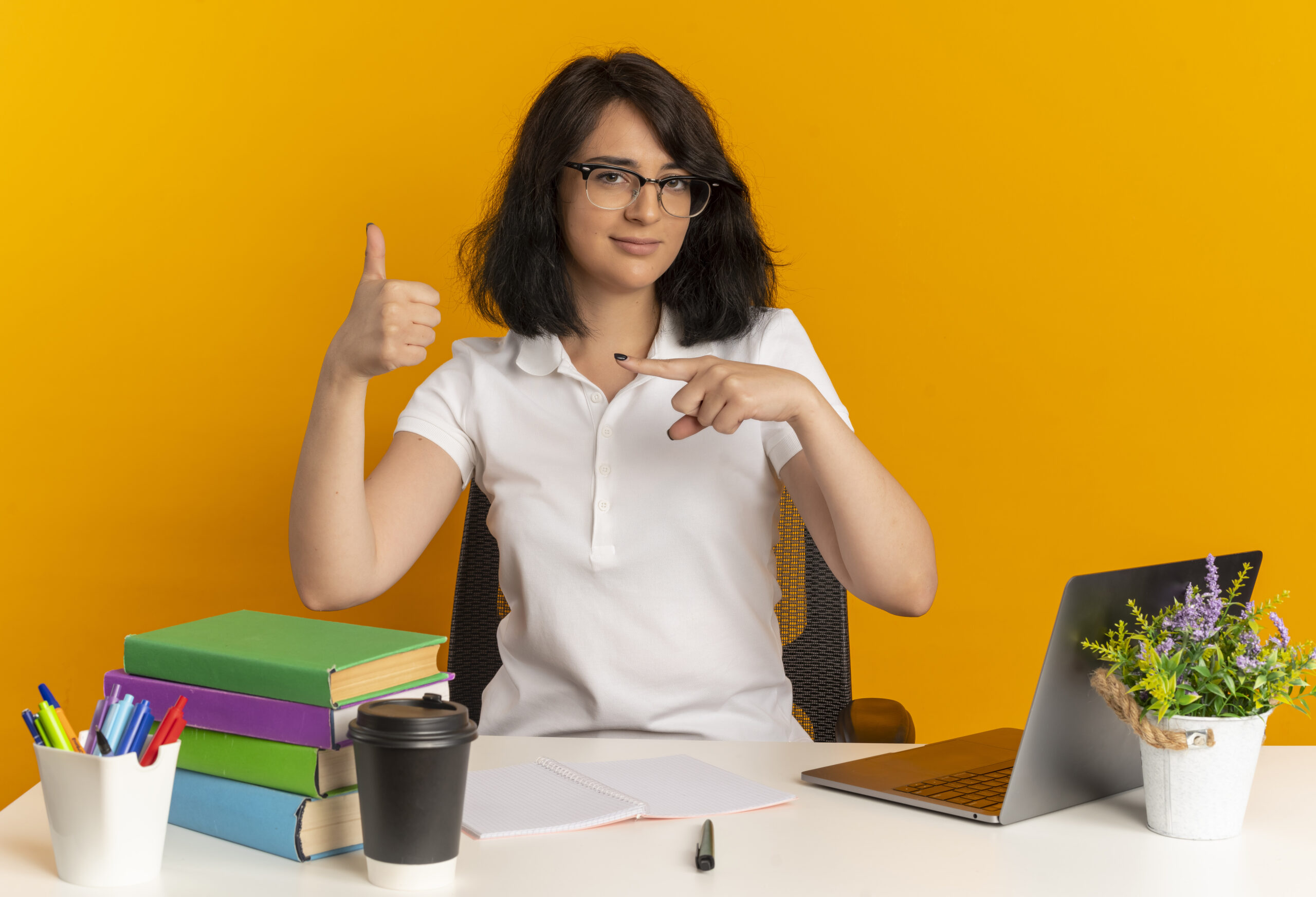 young confident pretty caucasian schoolgirl wearing glasses sits at desk with school tools thumbs up and points at side isolated on orange background with copy space